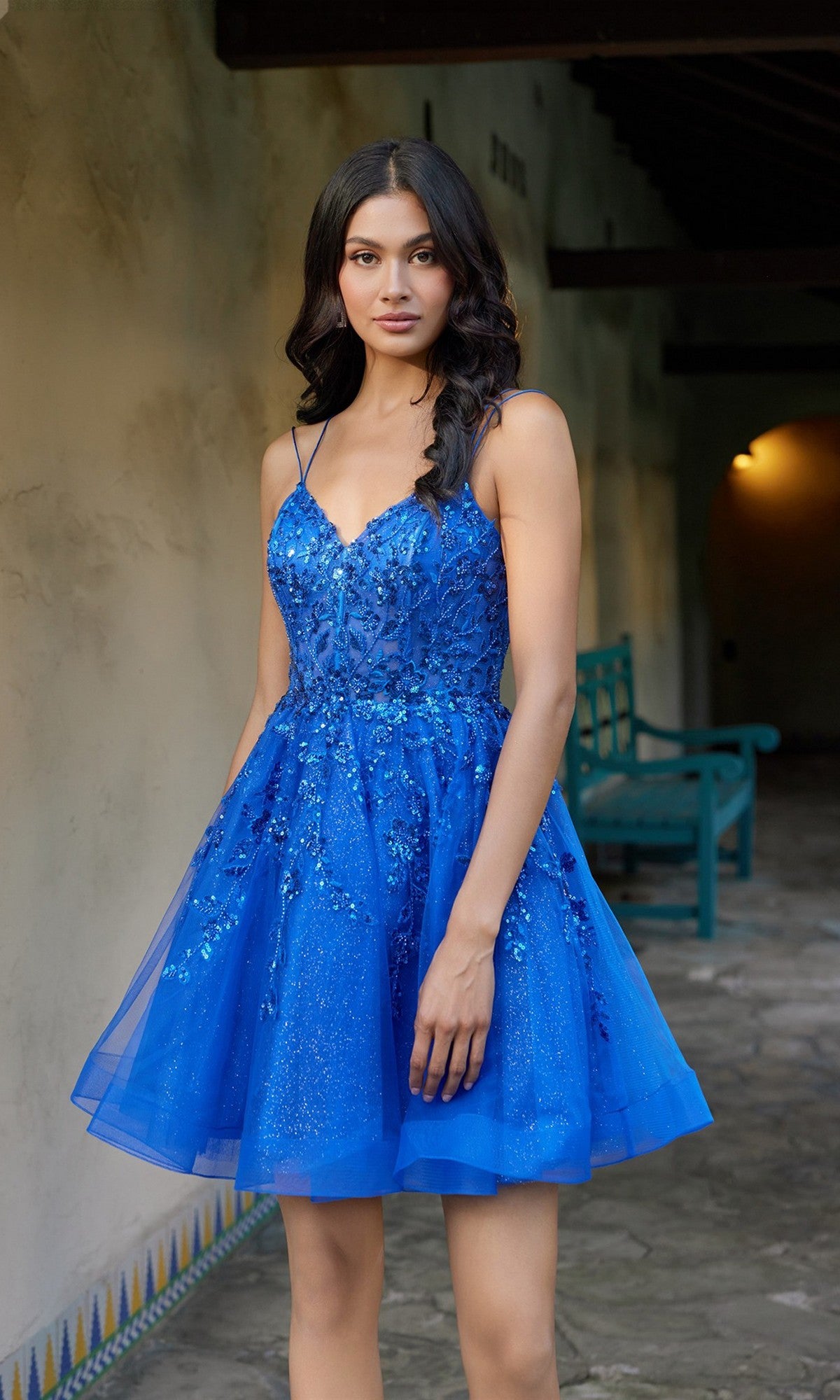 A woman wearing the Juliet JT988N short homecoming dress—a blue, sleeveless, fit-and-flare design with sparkly embellishments—stands on a stone walkway near a wall and a teal bench.